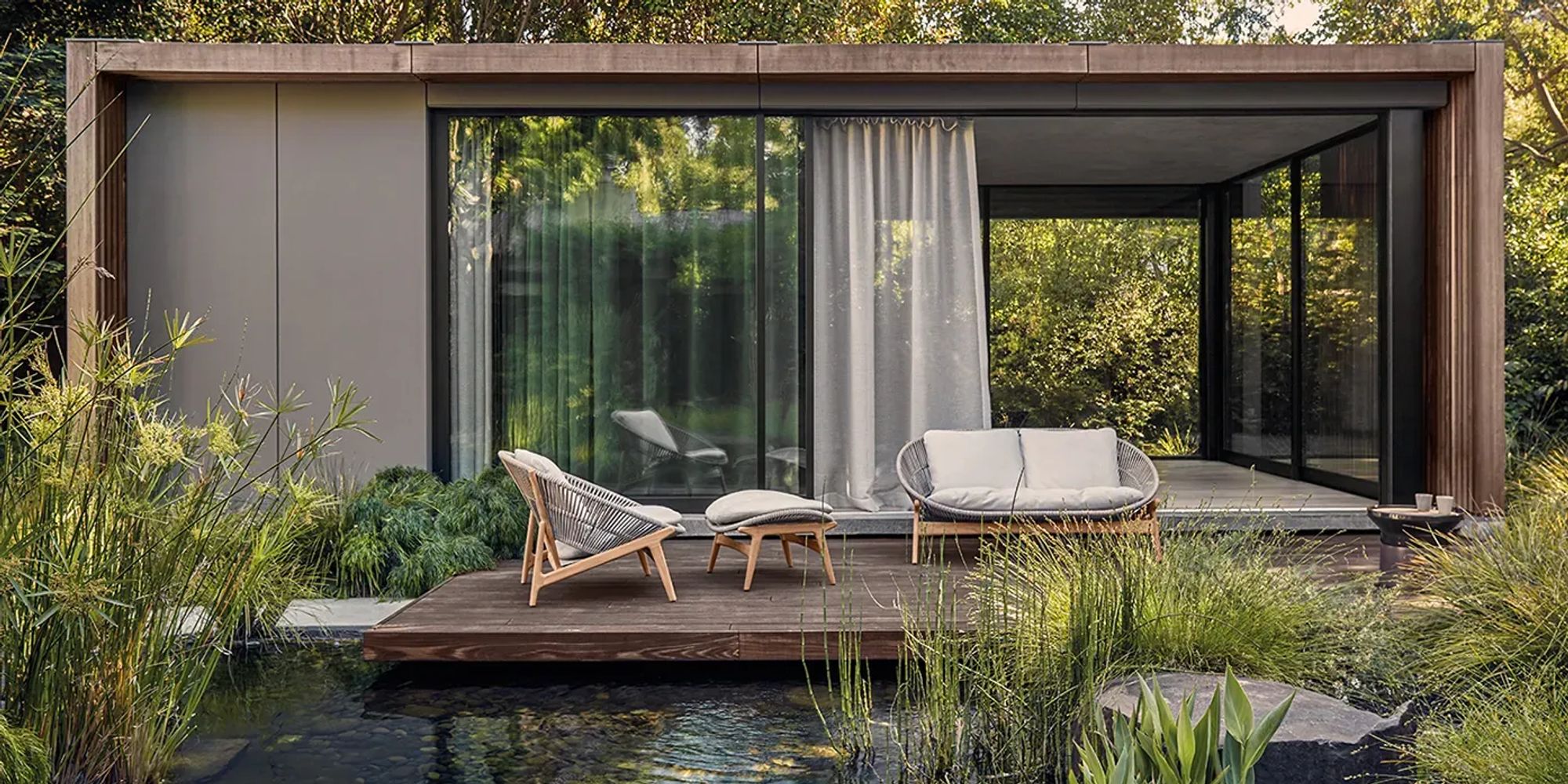 Gloster Bora lounge chairs and sofa on a timber deck beside a garden pond, modern pavilion backdrop, no people